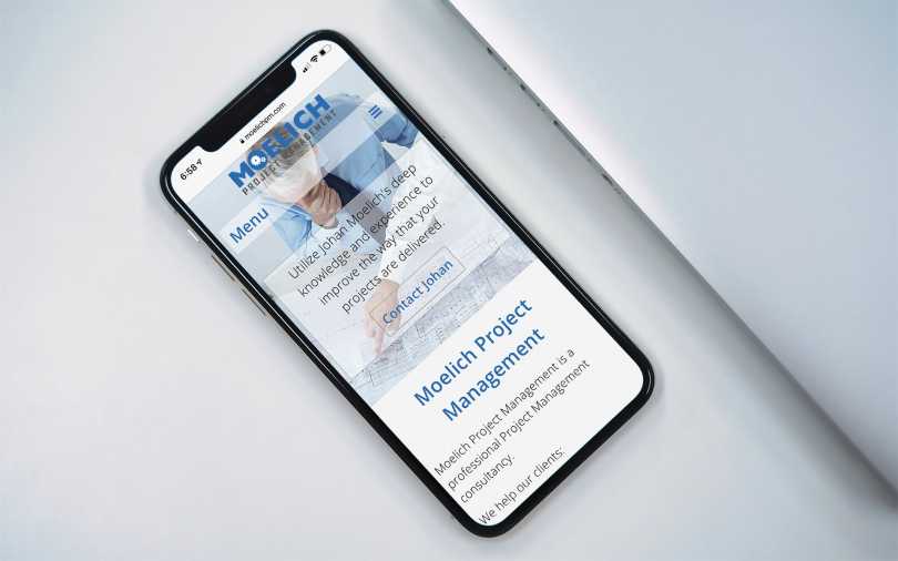 iPhone displaying Moelich Project Management home page, on table next to a laptop.