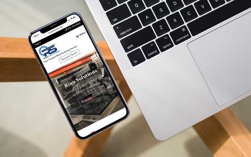 iPhone, displaying the Rian Solutions web site home page, sitting ona desk beside a laptop.