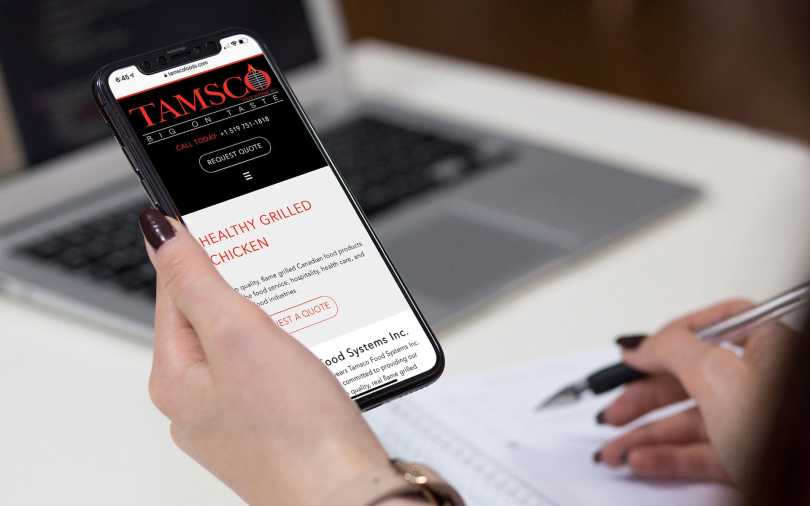 Tamsco Food Systems home page displayed on an iPhone being held by a woman.